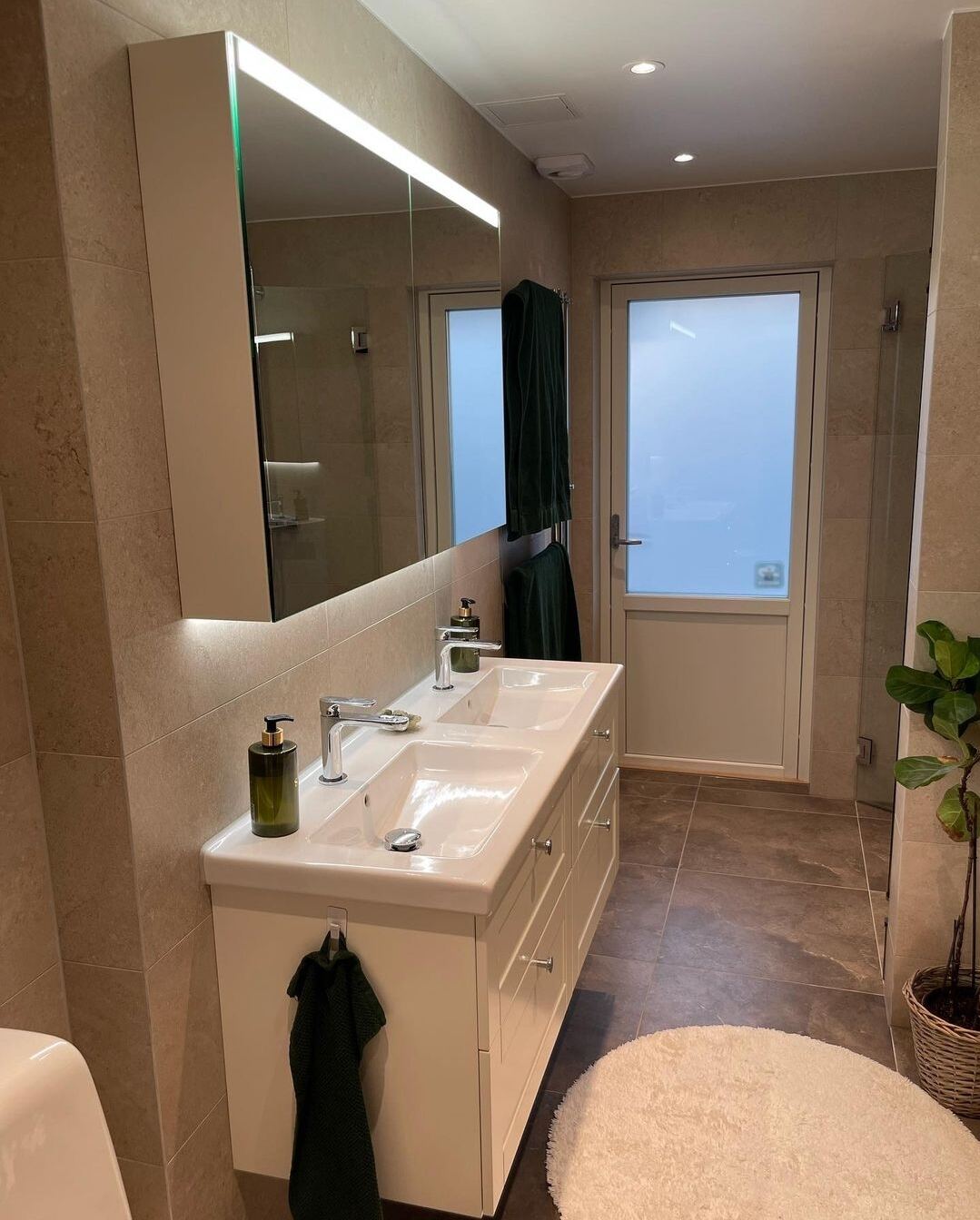 Renoverat badrum: Beige travertin-look, vit dubbelkommod, glasdusch, mörkt golv