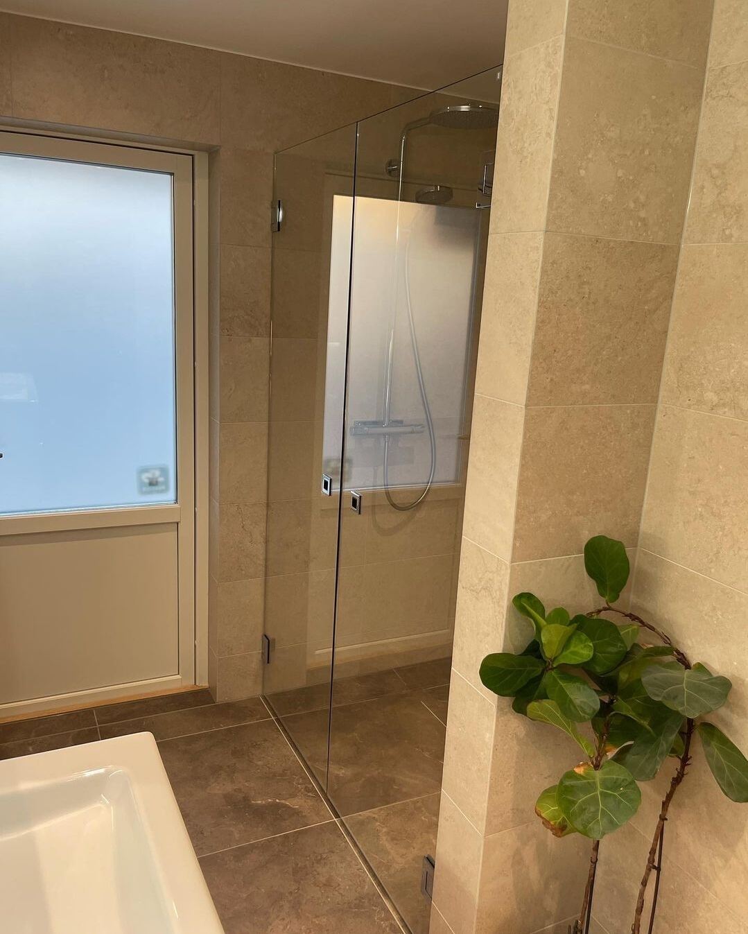 Renoverat badrum: Beige travertin-look, vit dubbelkommod, glasdusch, mörkt golv