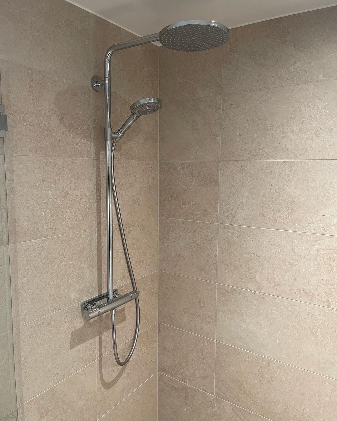 Renoverat badrum: Beige travertin-look, vit dubbelkommod, glasdusch, mörkt golv
